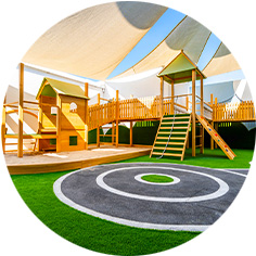 Outdoor playground with artificial turf, playhouses, a slide, climbing stairs, and shade sails overhead under a blue sky. Circular track marking visible in the foreground.