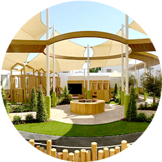 Outdoor playground with wooden structures, shaded sails, green artificial turf, plants, and circular seating area under a clear sky.
