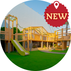 Outdoor modern wooden playground structure with slide and climbing areas, set on artificial grass. A red badge in the corner reads 