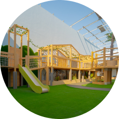 Outdoor playground with wooden climbing structures, slide, and artificial grass, enclosed by high walls and a partially open roof.
