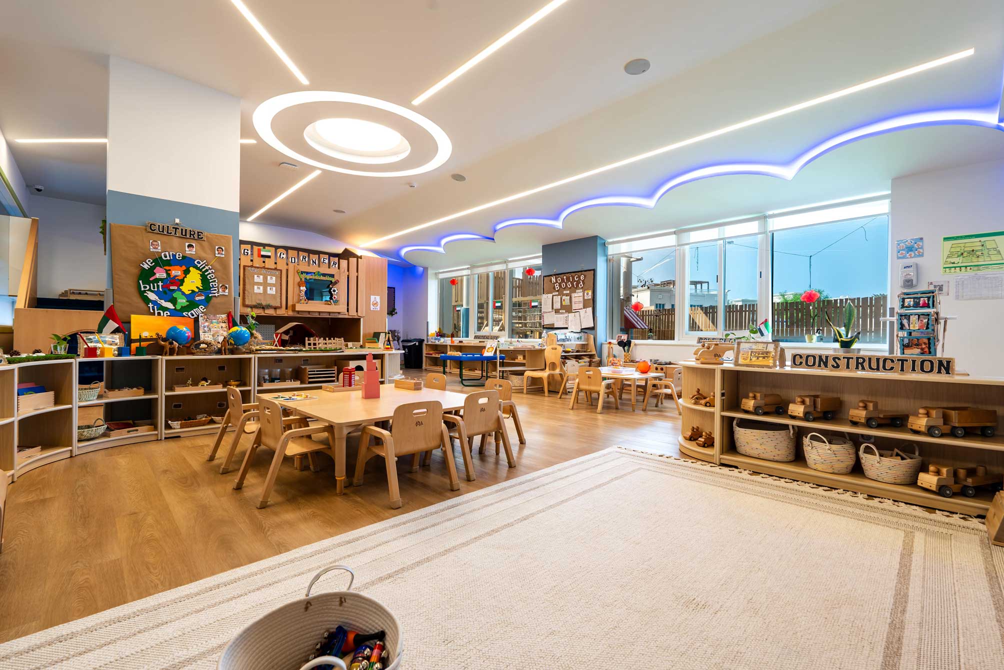 Bright, modern classroom with wooden furniture, low shelves displaying educational materials, a construction area, and colorful wall decorations. Large windows allow natural light in.
