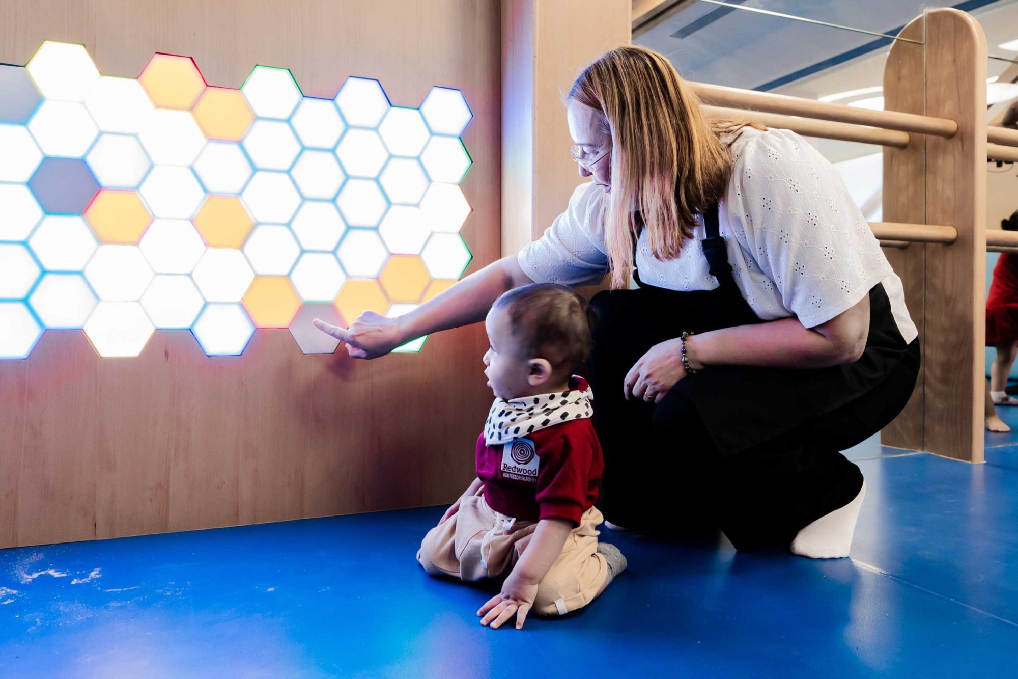 A woman and a child interact with a wall of illuminated hexagonal lights. The child, seated on a blue floor, reaches out to touch the lights as the woman assists.