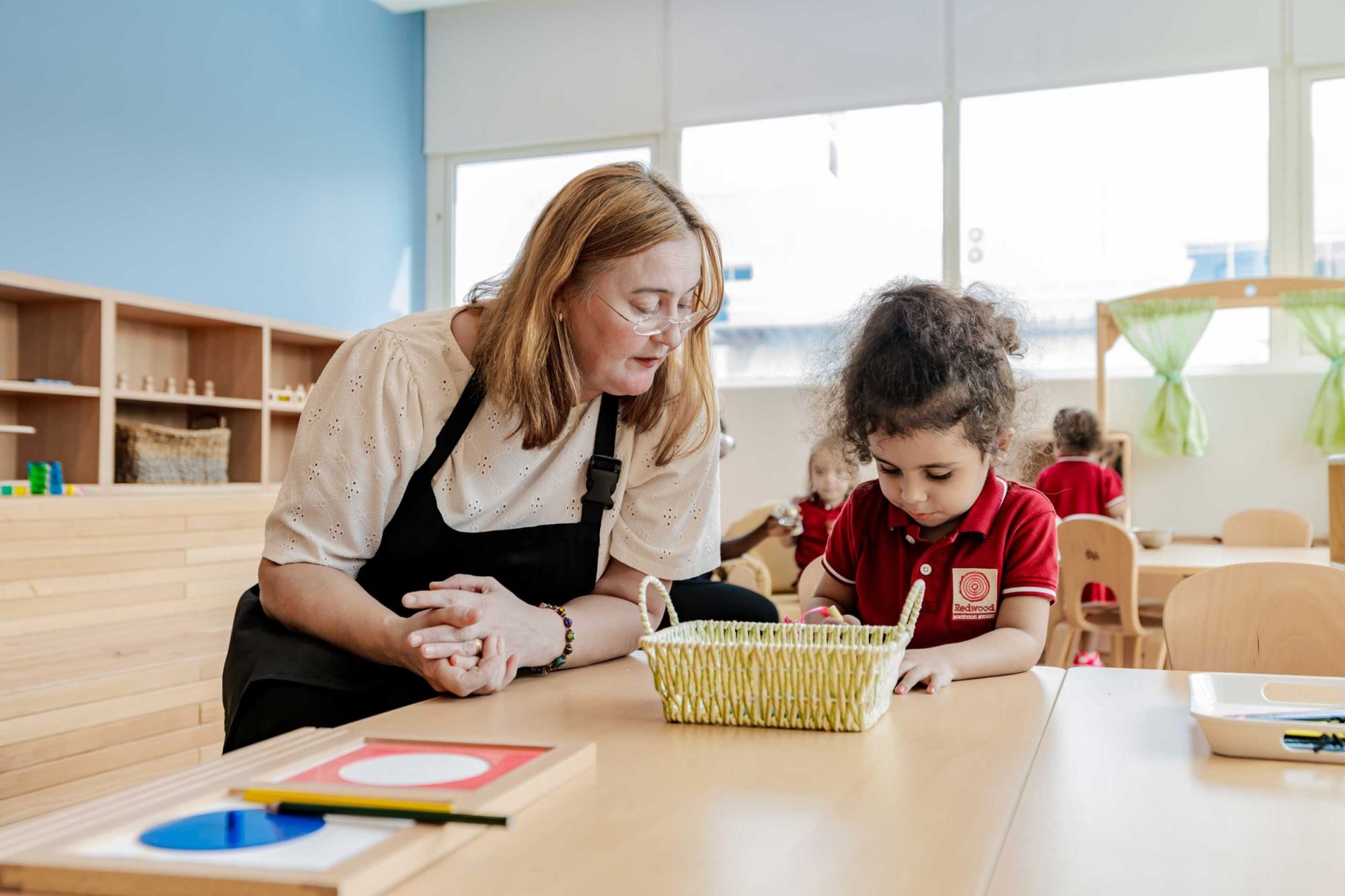 A teacher observes a young child working with a wooden puzzle at a classroom table, surrounded by wooden furniture and educational materials.