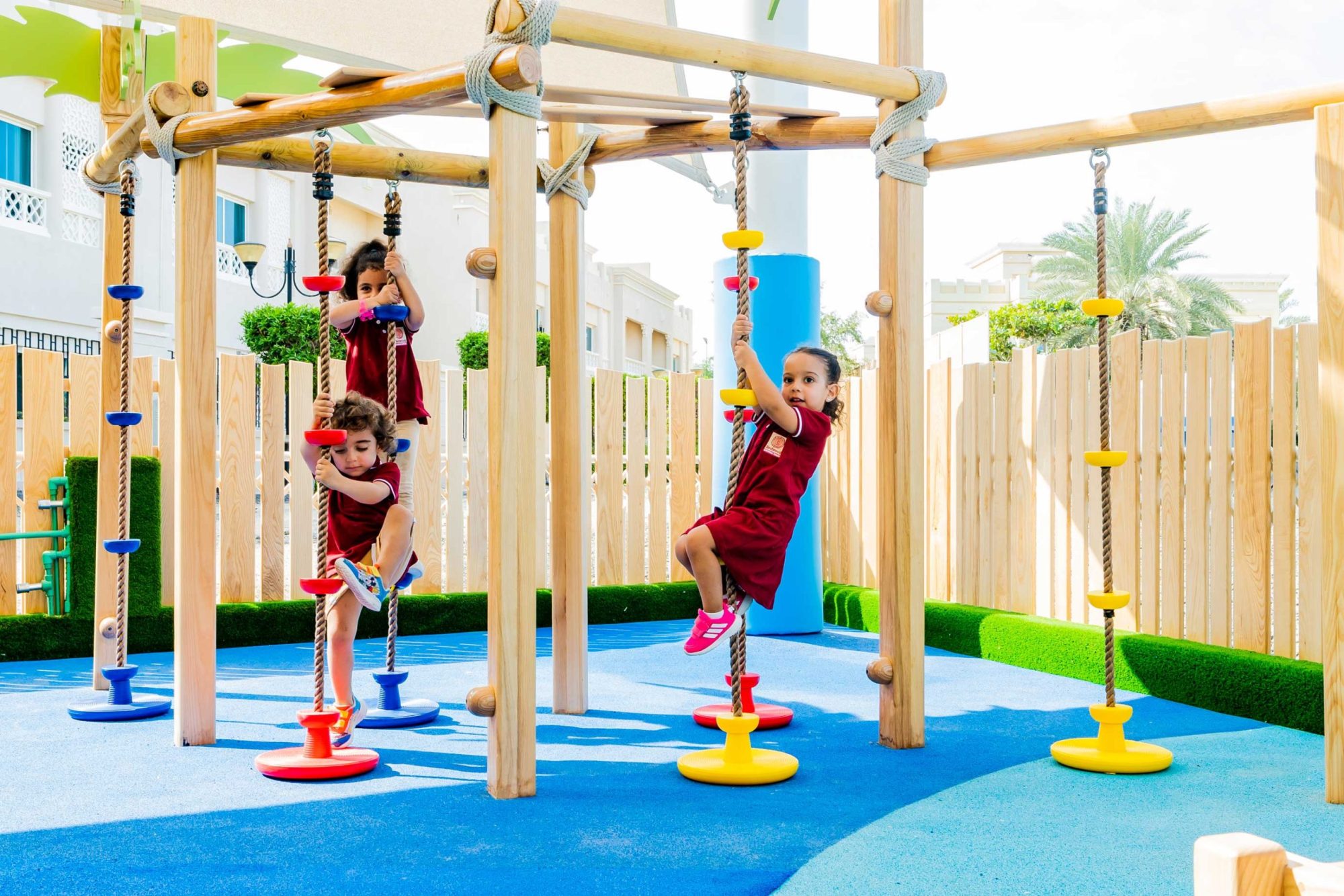 Three children in red uniforms play on rope and platform structures in an outdoor playground with a safety surface and wooden fence.