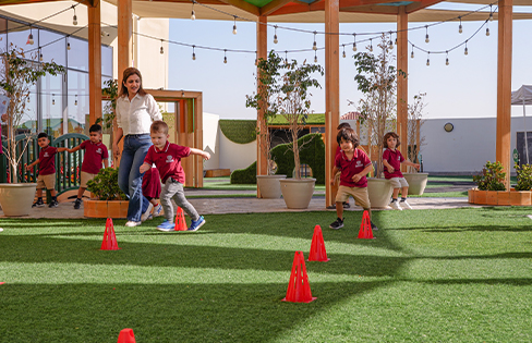 A group of young children and an adult run around red cones on a grassy outdoor playground under a shaded structure.