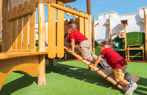 Three young children climb a wooden ramp on a playground structure outdoors on a sunny day.