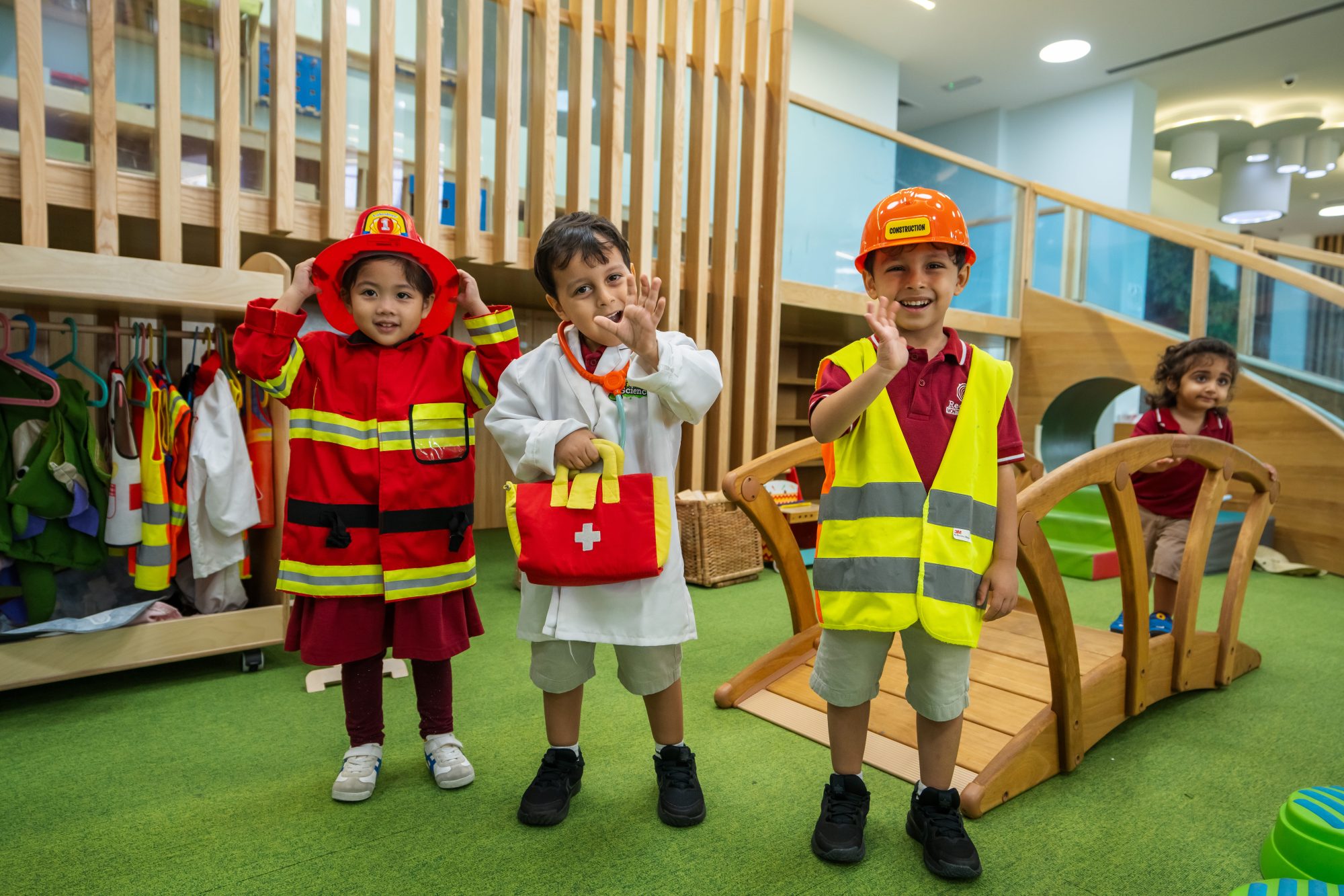 Three young children dressed in firefighter, doctor, and construction worker costumes stand and smile in a brightly lit playroom. Another child is visible in the background.