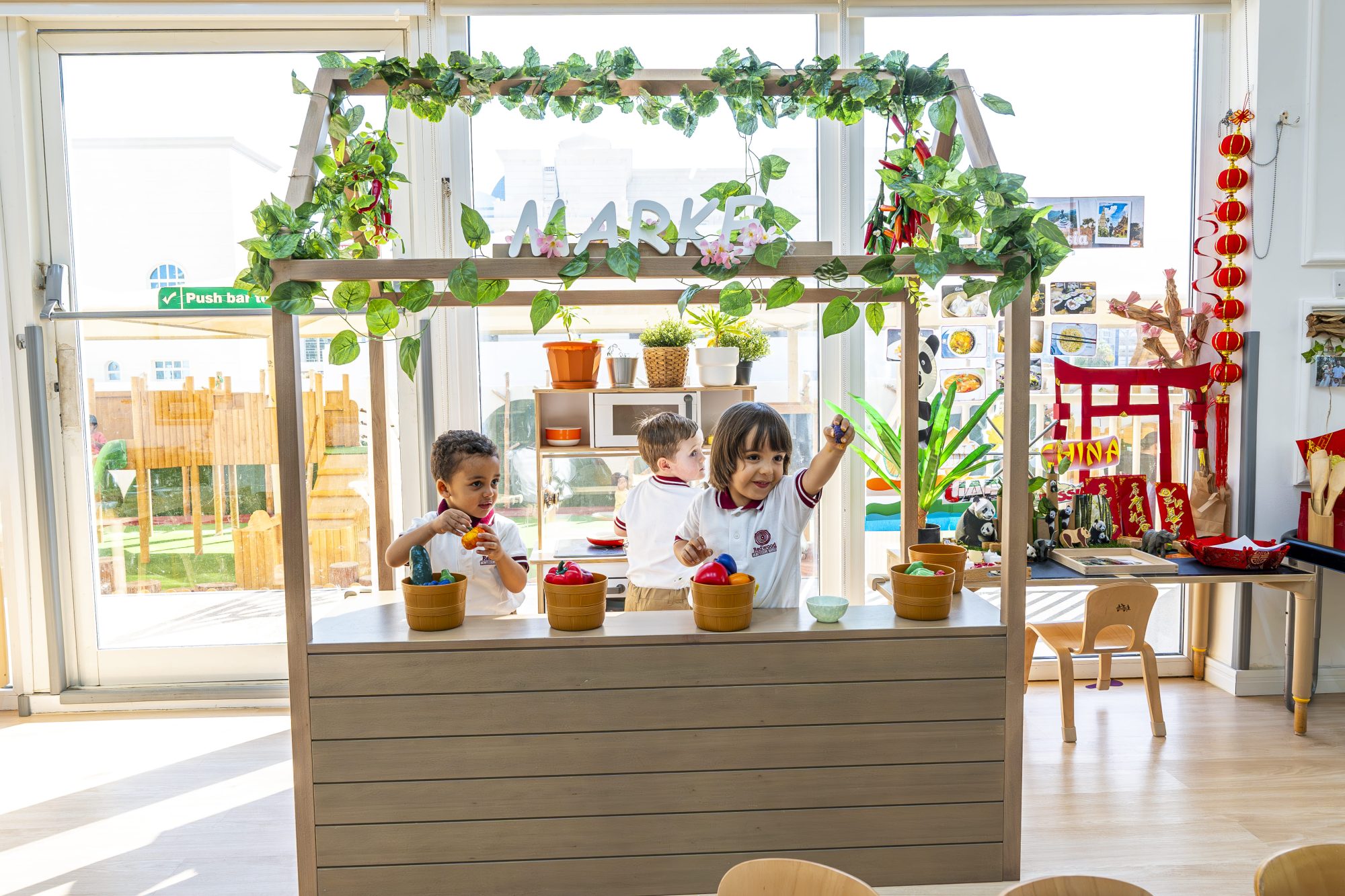Three children play with toy fruits and vegetables at a pretend market stand in a brightly lit classroom decorated with plants and festive ornaments.