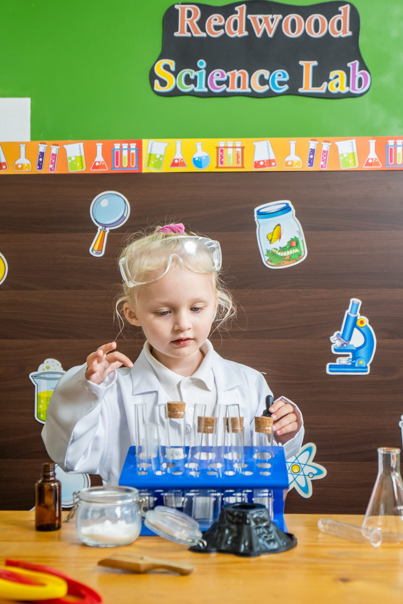 A young girl wearing safety goggles and a lab coat stands at a table with test tubes in a classroom labeled 