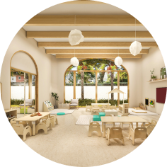 Bright, modern classroom with wooden furniture, play areas, large arched windows, and ceiling beams. Natural light fills the room, and outdoor scenery is visible through the windows.